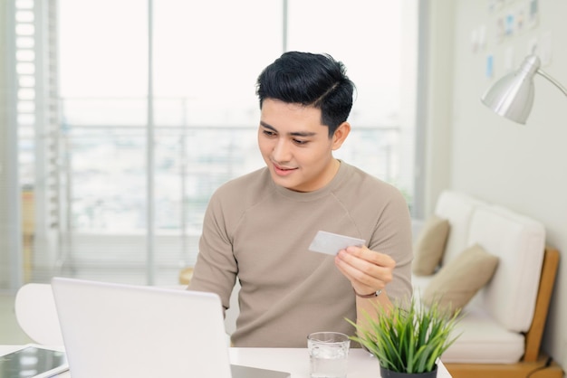 Apuesto joven empresario usando una computadora mientras sostiene una tarjeta de crédito en una oficina en casa