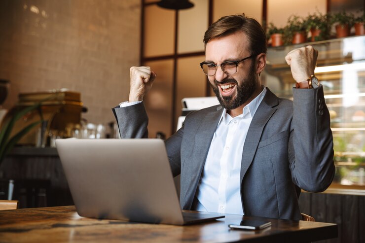 Foto apuesto joven empresario vestido con traje trabajando en una computadora portátil, sentado en el café en el interior, celebrando el éxito