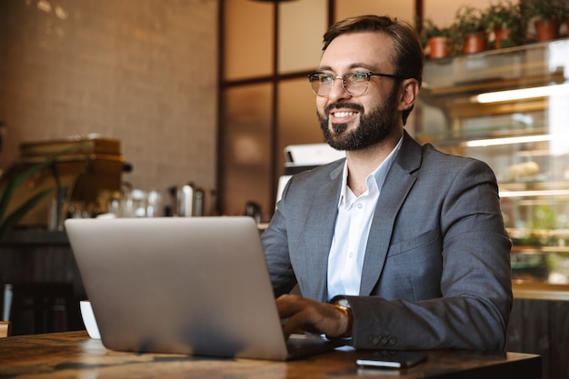 Apuesto joven empresario vestido con traje trabajando en una computadora portátil, sentado en el café en el interior, sosteniendo el teléfono móvil