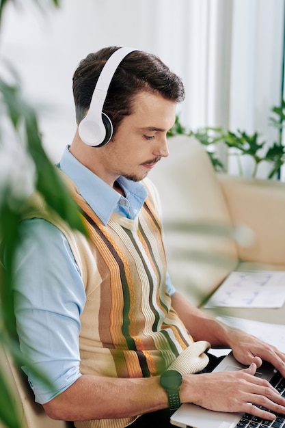 Apuesto joven escuchando música en auriculares cuando trabaja con una computadora portátil en la sala de estar de su apartamento