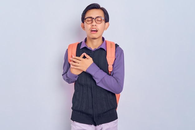 Apuesto joven estudiante asiático hombre vestido con ropa casual con mochila con las manos dobladas sobre su pecho corazón aislado sobre fondo gris concepto de colegio universitario de escuela secundaria