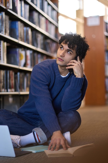 Apuesto joven estudiante latinoamericano hablando por teléfono móvil mientras estudia en el campus de la biblioteca