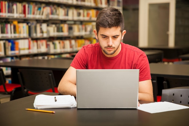 Apuesto joven estudiante universitario usando una computadora portátil para el trabajo escolar en la biblioteca