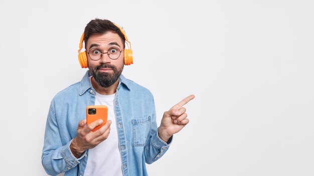 Apuesto joven europeo sorprendido con barba espesa escucha música de las redes sociales usa auriculares inalámbricos y el teléfono inteligente indica en el espacio de la copia contra la pared blanca