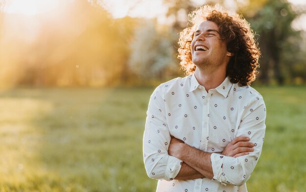 Apuesto joven feliz sonriendo con el pelo rizado posando en el parque de la ciudad mirando al cielo cruzando las manos y soñando Copiar espacio para su publicidad Concepto de estilo de vida y emoción de la gente