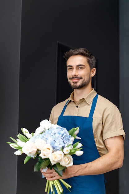 Apuesto joven florista masculino en delantal sosteniendo un hermoso ramo de flores y sonriendo a la cámara