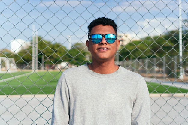 Apuesto joven con gafas de sol y suéter gris apoyado contra la valla de hierro