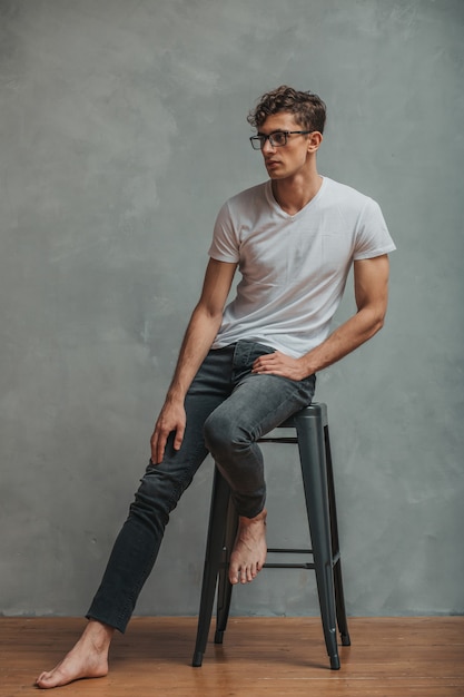 Apuesto joven con gafas, vestido con jeans y camiseta blanca posando silla descalza.