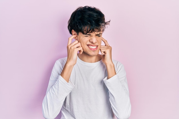 Apuesto joven hipster con camisa blanca casual que cubre las orejas con los dedos con expresión molesta por el ruido de la música a todo volumen concepto sordo