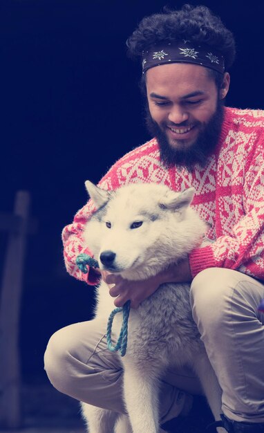 apuesto joven hipster jugando con un perro husky blanco frente a una vieja casa de madera retro vintage