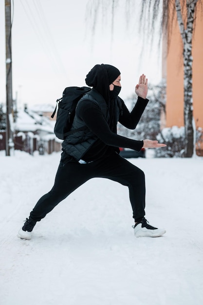 Un apuesto joven hipster con ropa negra de moda con una mochila y una máscara protectora posando afuera en un día de invierno. Genial chico escondido en un ninja de estilo negro cerca de la nieve.