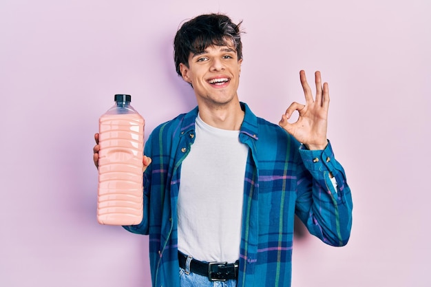 Apuesto joven hipster sosteniendo una botella de detergente haciendo el signo de ok con los dedos, sonriendo amigablemente gesticulando excelente símbolo