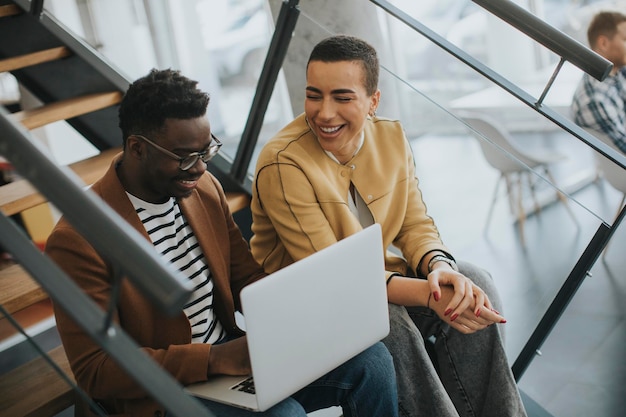 Apuesto joven hombre de negocios afroamericano y mujer de pelo corto trabajando en una laptop mientras se sienta en las escaleras de la oficina