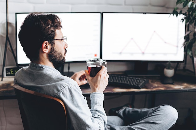 Un apuesto joven hombre de negocios con anteojos que trabaja con una computadora de forma remota sentado en una mesa de madera en casa Un hombre feliz y agradable que se comunica en las redes sociales buscando información en línea