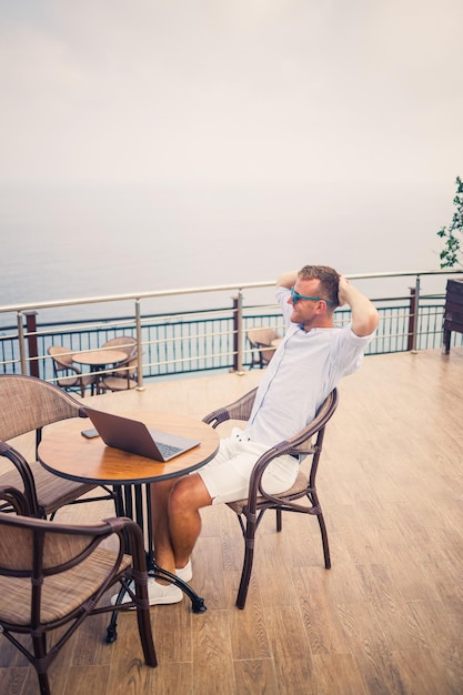 Apuesto joven hombre de negocios exitoso sentado en una mesa junto a la piscina con una computadora portátil con vista al mar Mediterráneo Trabajo remoto en vacaciones Concepto de vacaciones