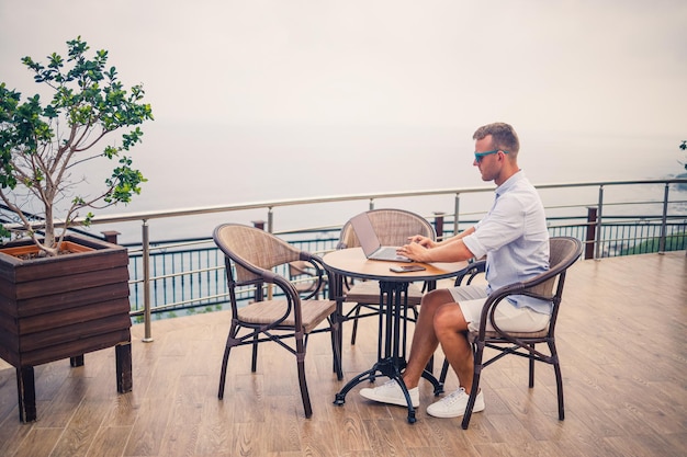 Apuesto joven hombre de negocios exitoso sentado en una mesa junto a la piscina con una computadora portátil con vista al mar Mediterráneo Trabajo remoto en vacaciones Concepto de vacaciones