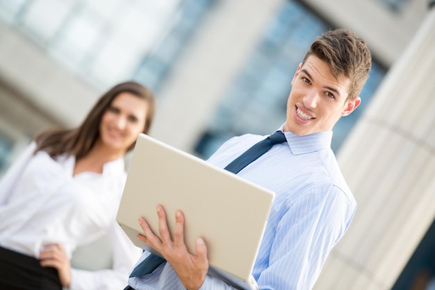 Un apuesto joven hombre de negocios con una laptop frente a su edificio de oficinas con su pareja en el fondo mirando la cámara.