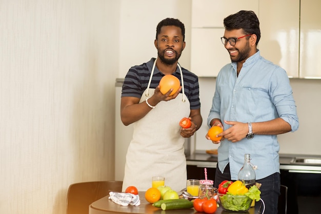 Apuesto joven indio con gafas y hombre afroamericano con delantal divirtiéndose en la cocina. Dos amigos varones de pie junto a la mesa con frutas y verduras, sonriendo haciendo batido. Copie el espacio.