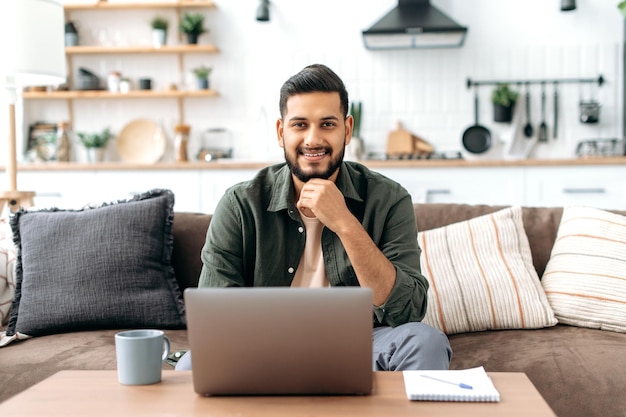 Un apuesto joven indio o árabe positivo y seguro de sí mismo vestido con ropa informal y elegante se sienta en casa en la sala de estar en el sofá con una laptop mirando a la cámara sonriendo amablemente