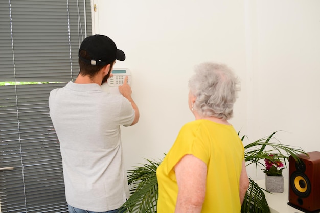 Apuesto joven instalando la seguridad de la casa cámara anti robo y alarma de sirena en una casa de mujer mayor