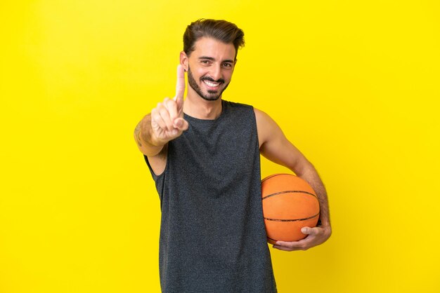 Apuesto joven jugador de baloncesto hombre aislado sobre fondo amarillo mostrando y levantando un dedo