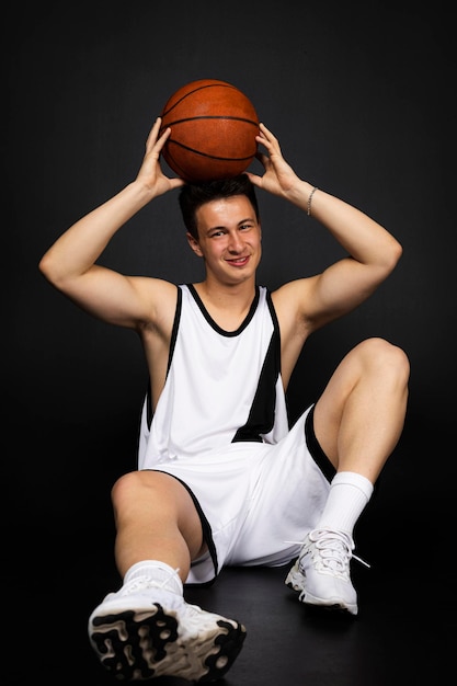 Apuesto joven jugador de baloncesto en ropa deportiva blanca se sienta en el suelo posando con una pelota aislado sobre fondo negro