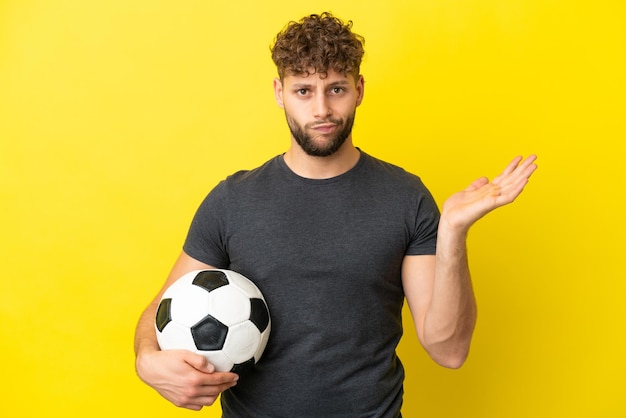Apuesto joven jugador de fútbol aislado de fondo amarillo que tiene dudas mientras levanta las manos