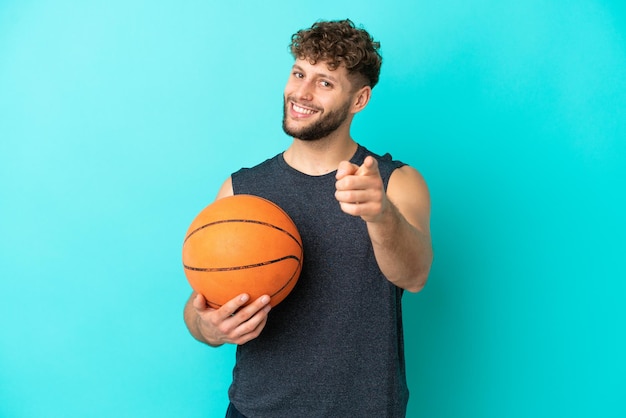 Apuesto joven jugando baloncesto aislado sobre fondo azul apuntando al frente con expresión feliz