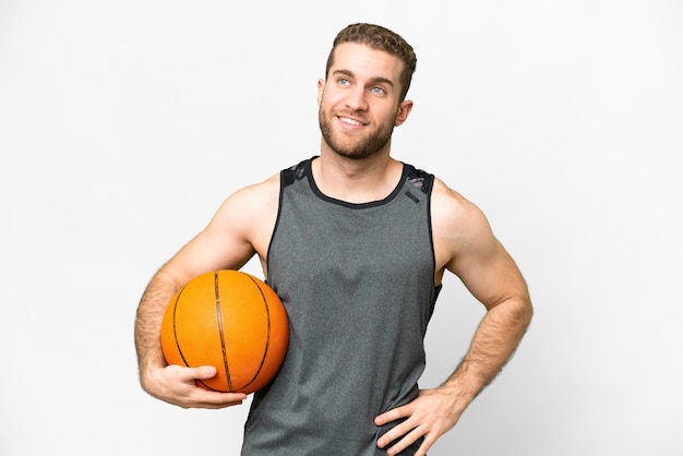 Apuesto joven jugando baloncesto sobre fondo blanco aislado posando con los brazos en la cadera y sonriendo