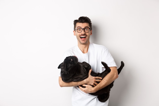 Apuesto joven jugando con lindo perrito negro. Dueño del perro acariciando a un pug, de pie sobre blanco