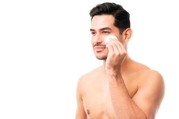 Apuesto joven limpiando su cara con una esponja facial sobre fondo blanco.