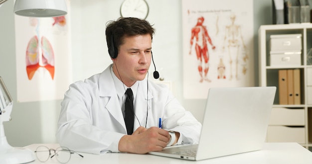 Un apuesto joven médico con auriculares delante de su portátil, hablando con un paciente.