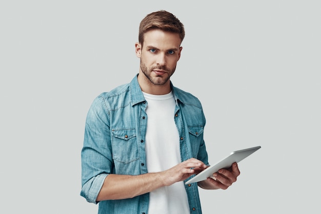 Apuesto joven mirando a cámara y usando tableta digital mientras está de pie contra el fondo gris