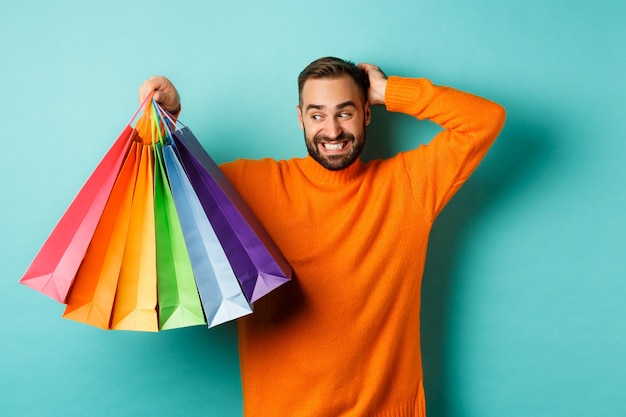 Apuesto joven mirando incómodo y rascándose la cabeza, mirando bolsas de compras con regalos, de pie sobre fondo turquesa.