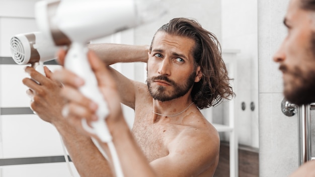 Apuesto joven mirándose en el espejo y secándose el cabello con un secador de pelo en casa