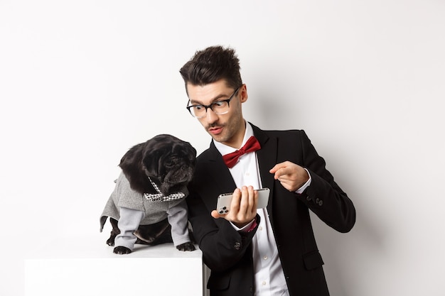 Foto apuesto joven mostrando algo en el teléfono móvil a su perro. propietario de compras en línea con mascota, de pie con disfraces sobre blanco.