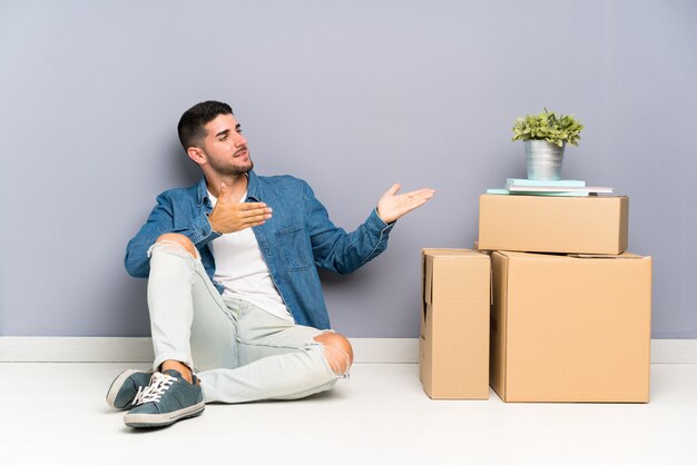 Apuesto joven moviéndose en una nueva casa entre cajas extendiendo las manos a un lado para invitar a venir