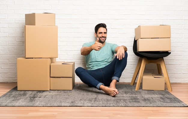 Apuesto joven moviéndose en un nuevo hogar entre cajas dando un gesto de pulgares arriba