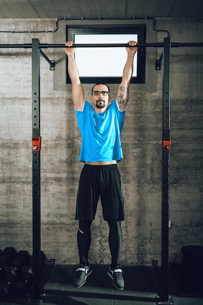 Un apuesto joven musculoso haciendo ejercicios de dominadas en el gimnasio. Mirando a la cámara.