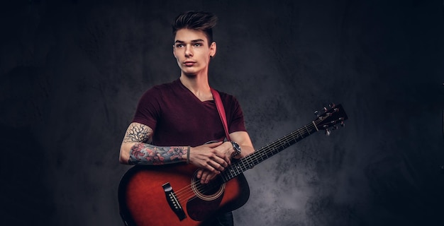 Apuesto joven músico con cabello elegante en una camiseta posando con una guitarra en sus manos sobre un fondo oscuro.