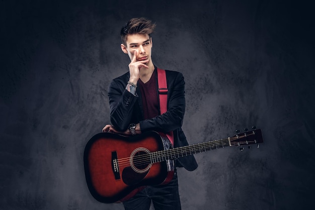 Apuesto joven músico pensativo con cabello elegante en ropa elegante posando con una guitarra en sus manos. Aislado en un fondo oscuro.