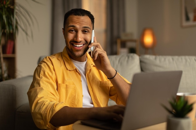 Apuesto joven negro con ropa informal hablando por teléfono inteligente disfrutando de una conversación en la oficina de casa