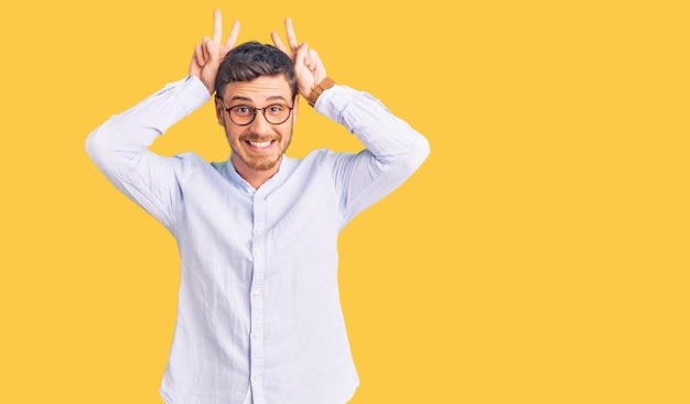 Apuesto joven con oso con elegante camisa de negocios y gafas posando divertido y loco con los dedos en la cabeza como orejas de conejo sonriendo alegre