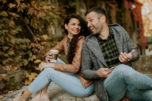 Apuesto joven pareja sentada en las escaleras al aire libre en un día de otoño y mediante teléfono móvil