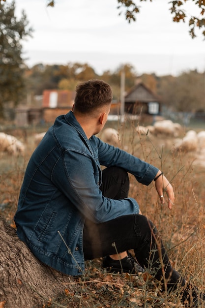 Apuesto joven con peinado en una camisa de mezclilla de moda y jeans se sienta y descansa cerca de un árbol en el campo y mira ovejas Guy viaja en el campo