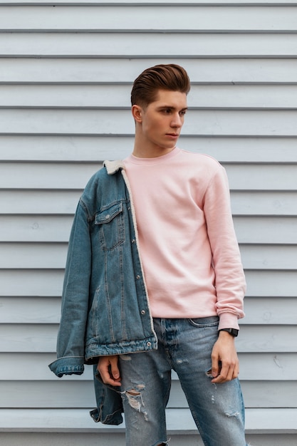 Apuesto joven con peinado con elegante chaqueta de mezclilla azul en sudadera de moda rosa junto a la pared blanca en la ciudad. Chico dulce urbano glamour en hermosos paseos de moda casual ropa. estilo callejero