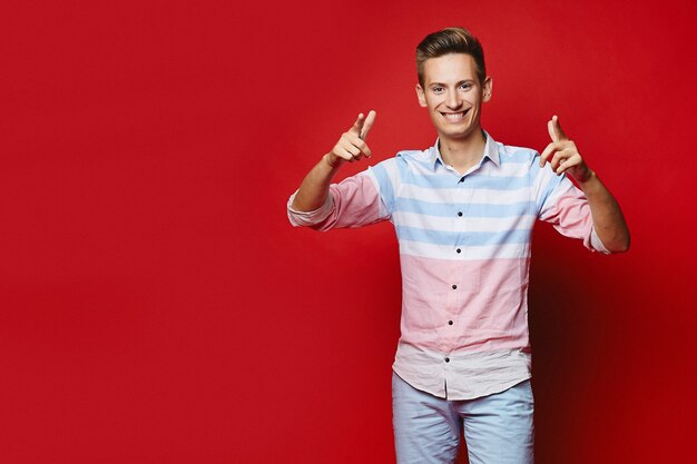 Apuesto joven con peinado de moda apunta a la cámara sobre un fondo rojo. Impresionante modelo masculino en camisa y pantalones cortos posando en el estudio