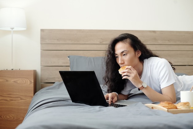 Apuesto joven con el pelo largo acostado en la cama comiendo bollo fresco y viendo un seminario web o un tutorial sobre ...