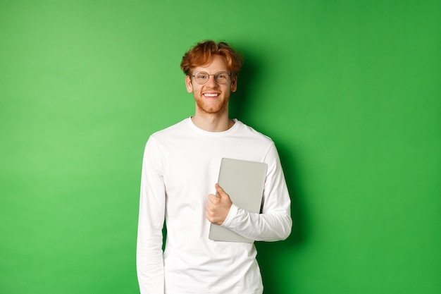 Apuesto joven con pelo rojo, gafas y camiseta de manga larga, sosteniendo portátil sobre fondo verde.