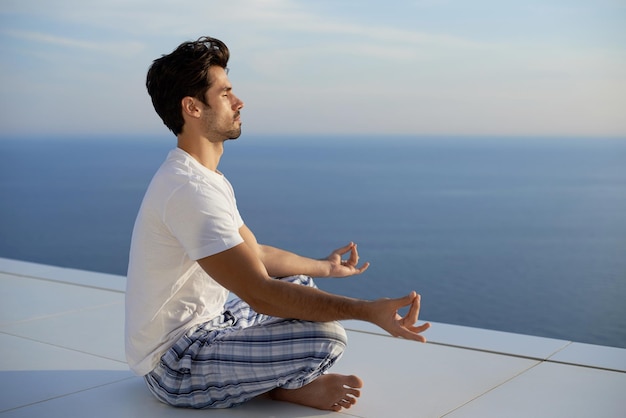 apuesto joven practicando yoga en la moderna terraza de la casa con el océano y la puesta de sol en el fondo